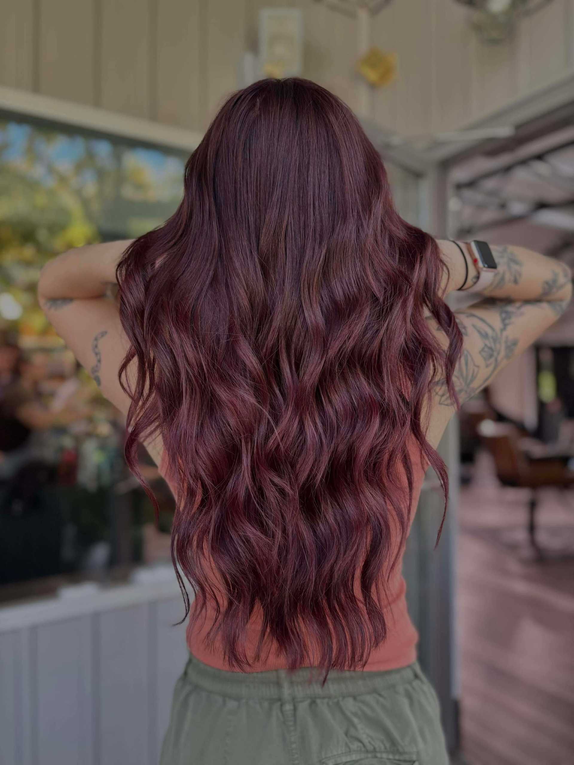 Woman with long wavy maroon hair stands in a casual setting, showing hair length and color.