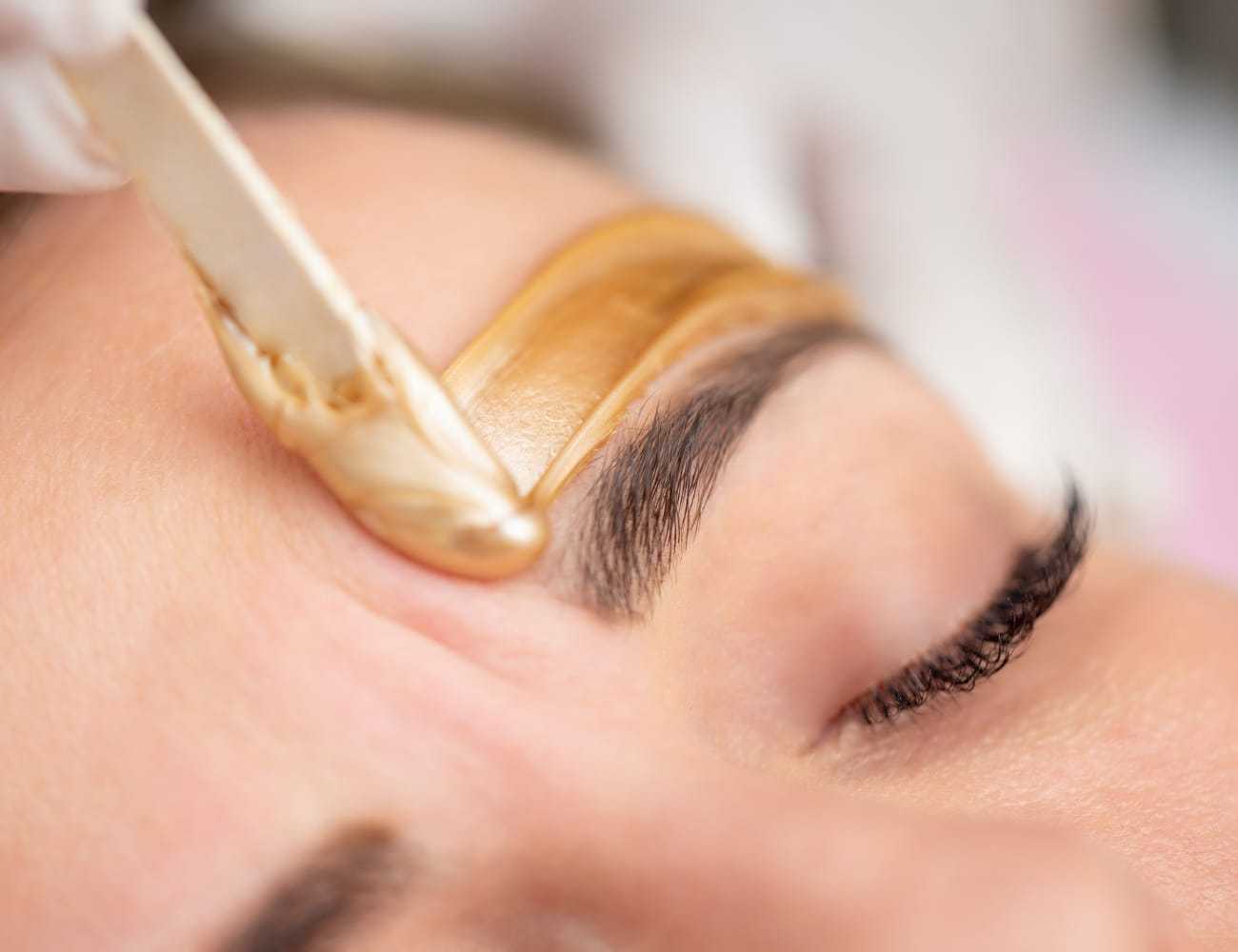 Eyebrow waxing with a wooden spatula applying warm wax on a person's face.
